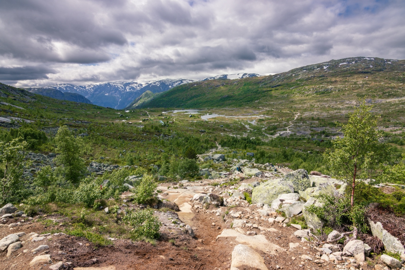 A rocky trail dropping into a green upland valley with small lakes and snow-streaked peaks under cloud