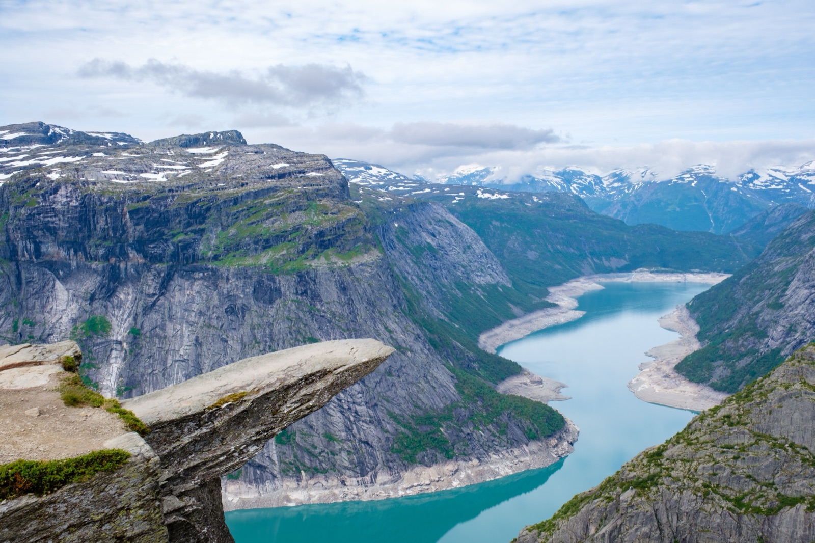 The Trolltunga rock ledge jutting out over a long turquoise lake between steep, snow-streaked mountains